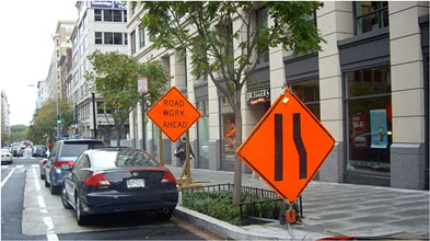 Photo of a lane ends symbol warning sign and a Road Work Ahead warning sign positioned on a sidewalk in an urban area.