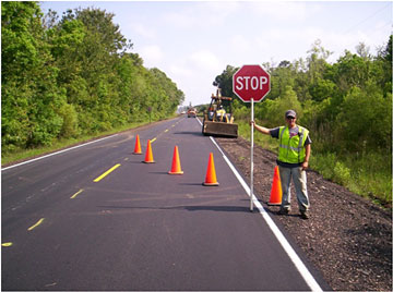 Photo of a flagger on a roadway shoulder next to the first cone in a lane taper holding a STOP sign and facing the direction of traffic.