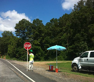 Photo of a flagger on a roadway shoulder holding a STOP sign but facing away from the direction of traffic.