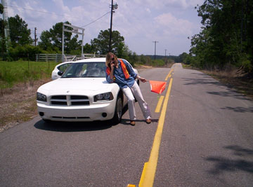 Photo of a flagger on a roadway leaning on a car hood and talking on a cell phone. The flagger is holding a flag but letting it hang down, is wearing a safety vest that is not fastened in front, and is not wearing a helmet.