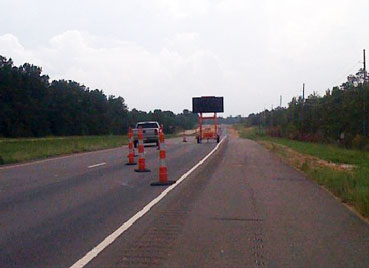Photo of four orange and white striped tubular markers on a roadway in a taper from the roadway shoulder to the broken white lane line and in advance of a portable changeable message sign.
