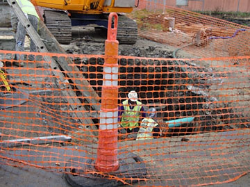 Photo of orange netting stretched across an orange and white striped tubular marker in front of a trench in a roadway with two workers in the bottom of the trench.