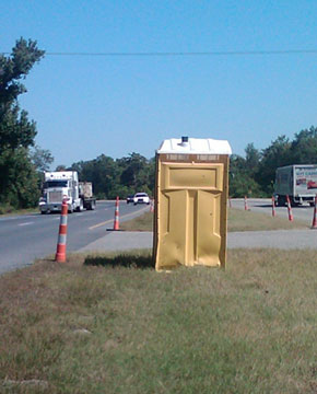 Photo of a dented portable toilet on a highway median with orange and white striped tubular markers on both sides of the median.