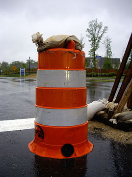 Photo of a dented orange and white striped drum on a roadway with a sandbag on its top.