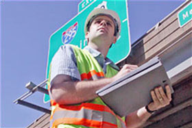 Photo of a man in a helmet and yellow-green safety vest holding a clipboard.