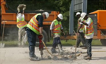 Photo of five workers in helmets and yellow-green safety vests using jack hammers in front of work vehicles on a roadway.