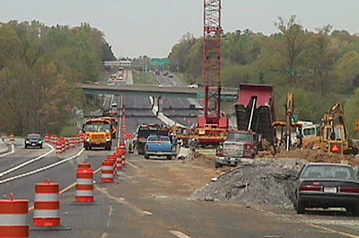 Photo of construction in a work zone across two lanes of a divided highway with orange and white striped drums separating the work zone from a traffic lane.