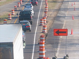 Photo of vehicles on a highway traveling in a single lane between orange and white striped drums.