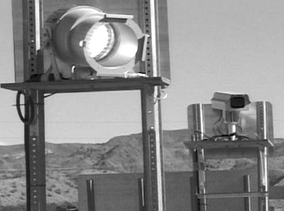 This figure shows a detailed view of one of the monitoring stations that was used to capture the license plate information of cars traveling through the work zone. The monitoring station consists of two small pole-mounted signs with a light mounted on the back of one sign, and a camera mounted on the back of the other. Mountains can be seen in the distance.