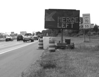 This figure shows the changeable message sign added to the system from an earlier study's recommendation. The sign shows an arrow pointed downward and to the left, with the text reading Merge Left. There are two drums with flashing lights in front of the trailer, and a speed limit sign is shown. The speed limit for I-94 is normally 70 miles per hour with a 55 mile per hour limit for trucks, as shown in the sign.