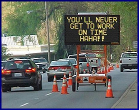 A humorous, photoshopped photo of heavy traffic passing moving slowly past a closed lane. Within the closed lane a changeable message sign reads 'You'll never get to work on time! Ha Ha!'